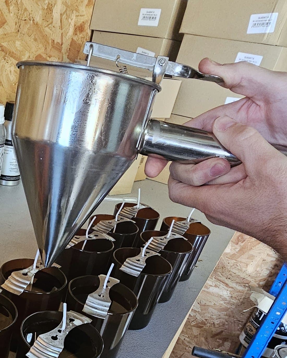 A hand pouring melted wax from a steel funnel into a row of amber jars, each wick held centred by a metal bar.