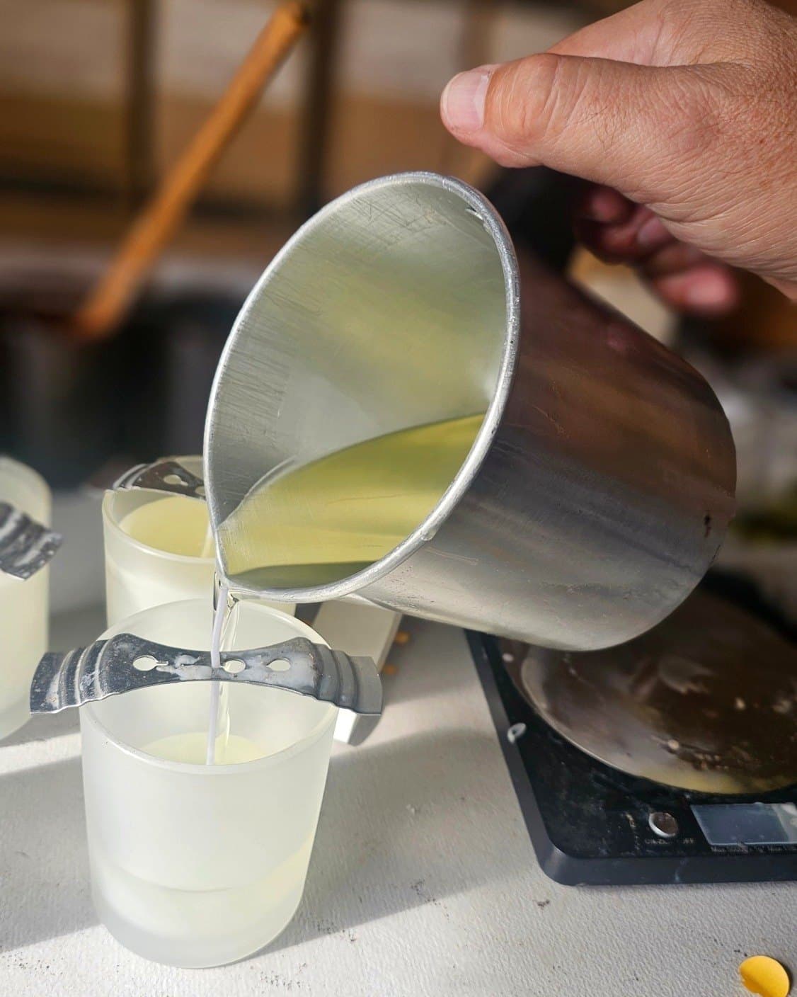 A hand tipping a steel jug of melted wax into a frosted glass jar, the wick held centred by a metal bar.