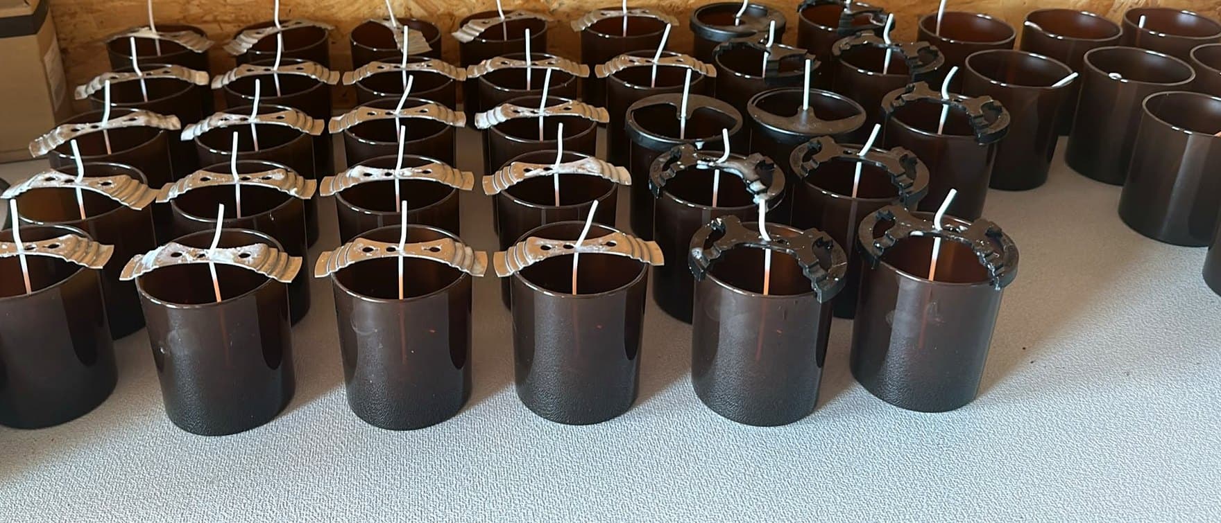Rows of empty amber and black candle jars on the studio bench, each wick held centred by a metal bar.